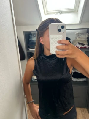 Svart ärmlös polotopp i satin - Snygg svart ärmlös polotopp i glansigt satinliknande material. Toppen har en hög krage och sitter tight mot kroppen. Enkel men ändå elegant look. Använd en gång så mycket bra skick. Kom privat för fler bilder i andra vinklar och pris kan diskuteras💗