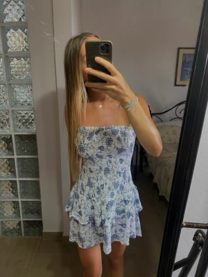Blommig ofshoulder klänning - Söt axelbandslös kortklänning i blått och vitt med blommigt mönster. Klänningen har volangdetaljer vid kjolen och smockad överdel för en bekväm passform. Perfekt för varma dagar och sommarens alla tillfällen. Xs men passar S,