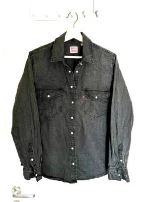 Svart jeansskjorta från Levi's - Säljer en svart jeansskjorta från Levi's med klassisk krage, vita tryckknappar och två bröstfickor med lock. Skjortan har lång ärm och en avslappnad passform. Perfekt för dig som gillar en tidlös och cool stil.