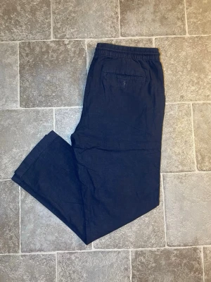 Mörkblå linnebyxor med dragsko - Snygga mörkblå linnebyxor med avslappnad passform och elastisk midja med dragsko. Byxorna har sidofickor och en bakficka, perfekta för en chill och stilren look. Tillverkade i lätt och luftigt linnematerial.