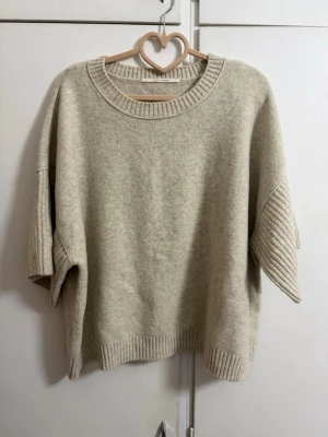 Beige oversized stickad tröja - Säljer en beige oversized stickad tröja med breda, halvlånga ärmar och ribbade detaljer vid hals, ärmslut och nederkant. Tröjan har en avslappnad passform och är gjord i ett mjukt material som passar perfekt för lager på lager.