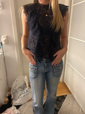 Marinblå spetsblus med volangdetaljer - Säljer en marinblå blus i spets med volangdetaljer längs framsidan och ärmhålen. Blusen är ärmlös och har en lätt transparent look som ger en trendig vibe. Perfekt att styla med jeans eller kjol för en cool outfit.