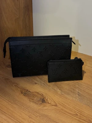 Svart axelväska monogram - Stilren svart axelväska i präglat läder med klassiskt monogrammönster. Väskan har dragkedja upptill, matchande liten plånbok och bred justerbar axelrem. Perfekt accessoar för dig som gillar lyxiga och trendiga detaljer.