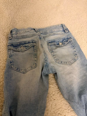 Ljusblå slitna jeans med knappar - Ljusblå jeans med slitningar på knäna och bakfickor med knappdetaljer. Jeansen har en klassisk femficksmodell och är tillverkade i denim. Perfekta för en avslappnad och trendig look.