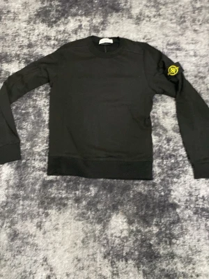 Svart sweatshirt från Stone Island - Svart sweatshirt från Stone Island med klassisk rund hals och ribbade muddar. På vänster ärm sitter det ikoniska Stone Island-märket i gult och grönt. Tröjan har en clean look och är tillverkad i mjukt bomullsmaterial.