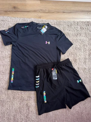 Svart träningsset från Under Armour - Säljer ett svart träningsset från Under Armour med t-shirt och shorts. Setet har färgglada logotyper och detaljer, samt elastisk midja på shortsen. Perfekt för gymmet eller löpning. Materialet är lätt och andas bra. Kan också kännas som mindre storlek eller större.