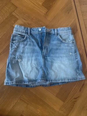 Ljusblå jeanskjol från Zara - Trendig ljusblå jeanskjol från Zara med slitna detaljer och klassisk femficksdesign. Kjolen har bälteshällor, knapp och dragkedja framtill. Perfekt för en avslappnad och cool stil. Materialet är denim i bomull.