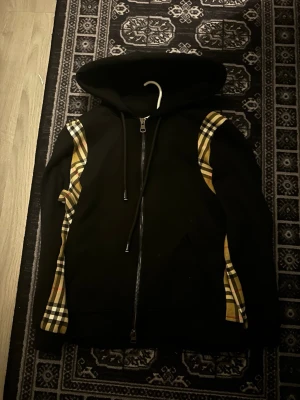 Burberry hoodie - Snygg svart hoodie från Burberry med klassiska beige-rutiga detaljer längs ärmarna. Hoodien har dragkedja framtill, justerbar huva med snören och mjukt material. Perfekt för dig som vill ha en stilren men ändå lyxig look.