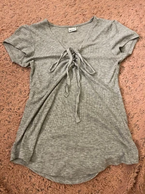 Grå ribbad topp med snörning - Snygg grå ribbad topp med korta ärmar och v-ringning. Toppen har en trendig snörning framtill som ger en cool detalj. Perfekt att styla med jeans eller kjol för en avslappnad look.