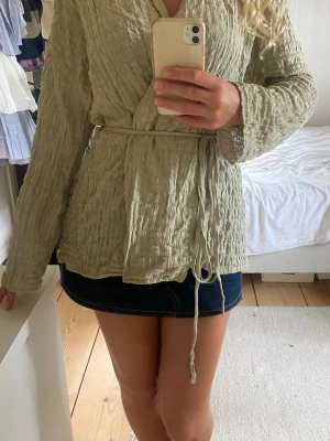 Beige omlottblus med knytskärp - Snygg beige omlottblus i ett krinklat tyg med knytskärp i midjan. Blusen har långa ärmar och en avslappnad passform som ger en chill vibe. Perfekt att styla med jeans eller kjol för en trendig look.