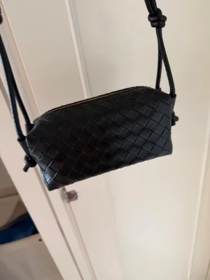 Svart flätad axelväska i läder - Snygg svart axelväska med flätad design och justerbar axelrem. Väskan är tillverkad i läder och har dragkedja upptill. Perfekt storlek för det viktigaste och ger en trendig touch till din outfit.