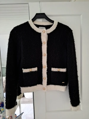 Svart kofta med vita detaljer och knappar - Stilren svart kofta med fluffig textur, vita kontrastkanter och dekorativa guldknappar framtill. Två fickor med vita detaljer på framsidan. Perfekt för dig som gillar klassisk men trendig look. Stl xs från Lindex. Den är nopprig men annars i bra skick!