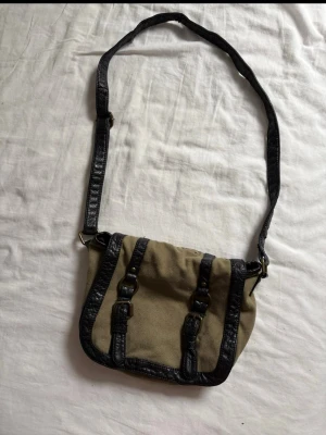 Beige axelväska med svarta detaljer - Snygg beige axelväska i canvas med svarta detaljer i fejkläder. Väskan har justerbar axelrem och dekorativa spännen framtill. Perfekt storlek för det viktigaste och ger en cool vintagekänsla till din outfit. Kolla gärna in min profil för bundle!
