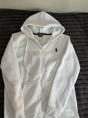 Vit hoodie från Polo Ralph Lauren - Snygg vit hoodie från Polo Ralph Lauren med dragkedja framtill och klassisk svart logga broderad på bröstet. Hoodien har huva med dragsko och är tillverkad i mjukt material för extra komfort. Perfekt för en avslappnad och stilren look.