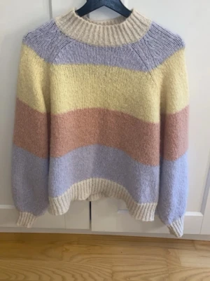 Pastellrandig stickad tröja - Mysig stickad tröja med breda pastellfärgade ränder i lila, gul, rosa och beige. Tröjan har en hög ribbad krage, ribbade muddar och avslut. Perfekt för dig som gillar en färgglad och avslappnad stil.