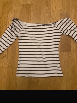 Randig offshoulder topp från F8 Sister - Säljer en vit offshoulder topp från F8 Sister med svarta horisontella ränder. Toppen har lång ärm och är figurnära i modellen. Perfekt för dig som gillar en enkel men snygg stil.