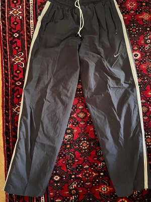 Svarta Nike mjukisbyxor med vita detaljer samt reflex på bägge sidorna - Snygga svarta/grå mjukisbyxor från Nike med vita ränder samt reflex längs sidorna och dragsko i midjan. Byxorna har raka ben, två fickor med dragkedja och en vit Nike-logga på vänster ben. Dragkedja nere på bägge ben. Perfekta för chill eller träning.