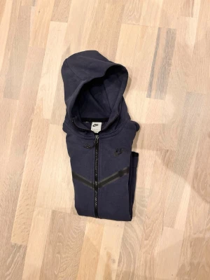 Mörkblå Nike tech - Snygg mörkblå hoodie från Nike med hel dragkedja framtill och huva. Hoodien har svarta detaljer över bröstet och en praktisk ficka med dragkedja på ena ärmen. Perfekt för en sportig och avslappnad stil.