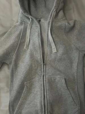 Ljusgrå zip hoodie russemerch - Ljusgrå hoodie med dragkedja från russemerch Klassisk modell med huva och vita dragsnören, samt kängurufickor framtill. Tillverkad i mjukt sweatshirtmaterial och ribbade muddar vid ärmslut och nederkant. Perfekt för en avslappnad stil.