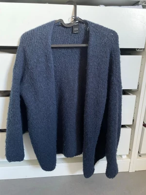 Mörkblå stickad kofta från H&M - Mysig mörkblå stickad kofta från H&M med öppen front och lång ärm. Koftan har en fluffig struktur och är perfekt att slänga på sig när det är kyligt. Enkel och stilren design som passar till det mesta.