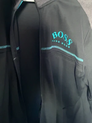  ziptröja från Hugo Boss - Svart ziptröja från Hugo Boss med turkosa detaljer och logga på bröstet. Tröjan har hel dragkedja, ribbade muddar och en sportig look. Tillverkad i mjukt material som känns skönt mot huden. Perfekt för en avslappnad och stilren outfit.