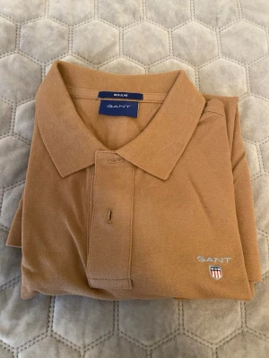 Beige pikétröja från Gant - Säljer en klassisk beige pikétröja från Gant med korta ärmar och krage. Tröjan har knappar framtill och Gant-logga broderad på bröstet. Perfekt till en avslappnad men ändå stilren look.