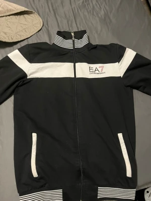 Svart ziptröja från Emporio Armani - Snygg svart ziptröja från Emporio Armani EA7 med vita detaljer och logga på bröstet. Tröjan har ståkrage med randig mudd, vita ränder över bröstet och på fickorna samt ribbade muddar vid ärmslut och nederkant. Perfekt till streetwear-looken.