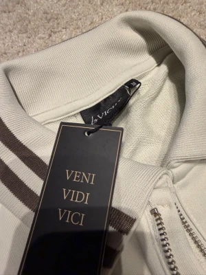 Beige ziptröja från La Vichy - Snygg beige ziptröja från La Vichy med bruna ränder på kragen och ärmsluten. Tröjan har hög krage, dragkedja framtill och är tillverkad i ett mjukt, stickat material. Perfekt för en avslappnad och trendig look.