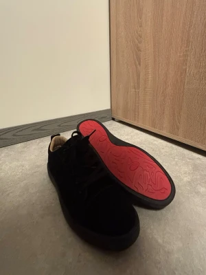 Svarta sneakers med röd sula - Säljer ett par stilrena svarta sneakers med snygg röd sula och klassisk snörning. Ovandelen är i mocka för en mjuk känsla och modern look. Perfekta för dig som vill ha något enkelt men ändå sticker ut med färgdetaljen under.