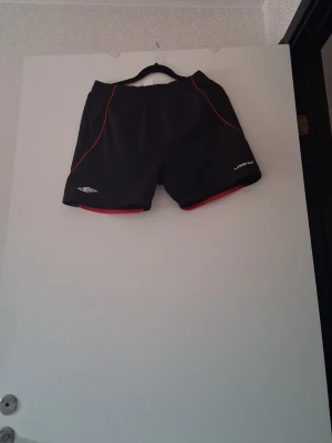 Svarta träningsshorts med röda detaljer - Svarta träningsshorts med röda sömmar och detaljer från Umbro. Shortsen har elastisk midja och är tillverkade i ett lätt och syntetiskt material som passar perfekt för sport och aktivitet.