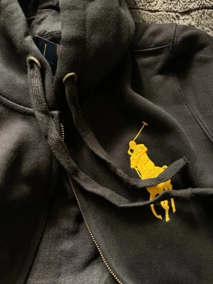 Svart hoodie med gul Polo-logga - Snygg svart hoodie med dragkedja från Polo Ralph Lauren. Stor gul broderad logga på bröstet och siffran 3 på ena ärmen. Mjuk bomull, huva med snören och ribbade muddar. Perfekt för en avslappnad och sportig stil.