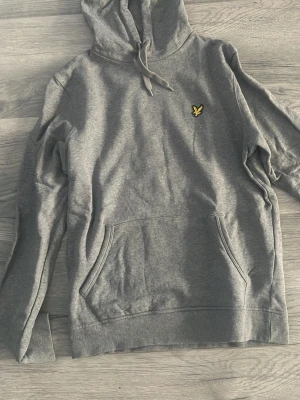 Grå hoodie med gul logga - Snygg grå hoodie med huva och dragsko samt en gul broderad logga på bröstet. Hoodien har en stor magficka och ribbade muddar vid ärmslut och nederkant. Perfekt för en avslappnad och stilren look.