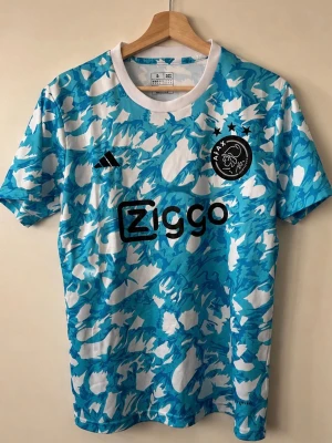 Ajax blåvit matchtröja med mönster - Säljer en Ajax matchtröja från Adidas i blått och vitt med ett unikt abstrakt mönster. Tröjan har korta ärmar, rund hals och Ajax-logga samt Ziggo-tryck på bröstet. Perfekt för fotbollsträning eller att bära till vardags.