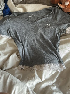 Grå ribbad topp från Hollister - Grå ribbad vintage topp från Hollister med v-ringning och spetsdetaljer vid ärmslut och nederkant. Kortärmad och figurnära passform, perfekt för en chill och snygg look. Diskret Hollister-logga broderad på bröstet.