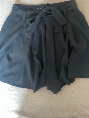 Svarta shorts med knyt och bälte - Säljer ett par svarta shorts med hög midja och ett brett knytband i midjan. Shortsen har en lös passform, sidofickor och en dekorativ bakficka. Perfekta för varma dagar och ger en stilren look.
