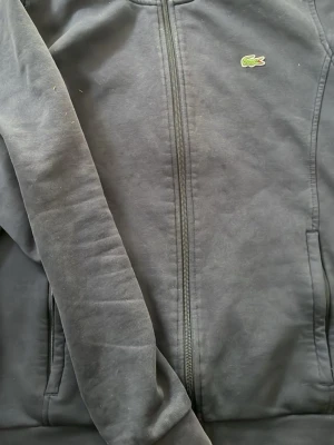 Mörkblå hoodie från Lacoste Sport - Snygg mörkblå hoodie från Lacoste Sport med dragkedja framtill och klassisk krokodillogga på bröstet. Tillverkad i mjukt material med fleece på insidan och ribbade muddar. Perfekt för en sportig och avslappnad stil.