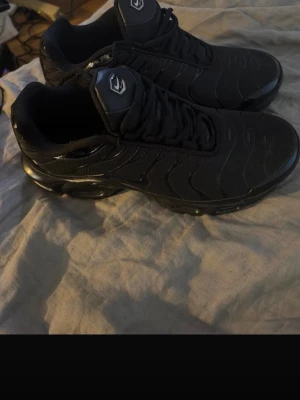 Svarta Nike Air Max Plus TN sneakers - Säljer ett par helsvarta Nike Air Max Plus TN sneakers med klassisk TN-logga bak. Skorna har en sportig siluett, mesh och syntetmaterial samt tydliga vågformade detaljer på ovandelen. Perfekta för dig som gillar streetwear och en clean look.