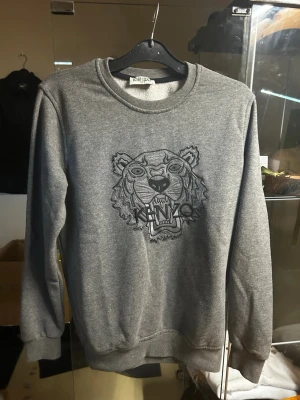 Grå sweatshirt från Kenzo med tiger  - Säljer en grå sweatshirt från Kenzo med det ikoniska tigermotivet broderat på bröstet. Tröjan har rund halsringning, ribbade muddar och är gjord i mjukt sweatshirtmaterial. Perfekt för dig som gillar streetwear och snygga detaljer.