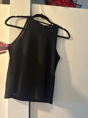 Svart ribbad ärmlös linnetopp - Snygg svart ärmlös linnetopp med ribbad struktur. Toppen har en hög halsringning och är tillverkad i ett lätt, tunt material som ger en stilren look. Perfekt att matcha med jeans eller kjol för en trendig outfit.