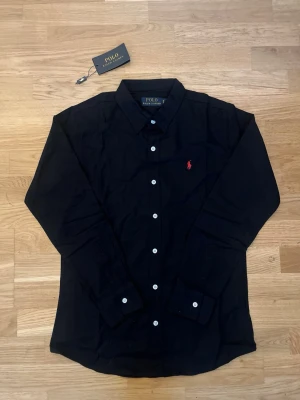 Svart skjorta från Polo Ralph Lauren - Klassisk svart skjorta från Polo Ralph Lauren med vit knappstängning och röd broderad logga på bröstet. Skjortan har lång ärm och normal passform, perfekt till jeans eller chinos. Tidlös design med krage och mjukt bomullsmaterial. Knappast änvänd, är storlek M men sitter mer mot S. Hör av dig vid frågor.