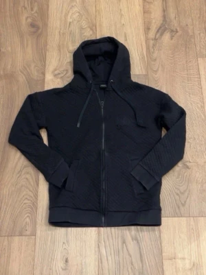 Svart hoodie från Hugo Boss (M) - Svart hoodie från Hugo Boss med dragkedja framtill och broderad logga på bröstet. Hoodien har quiltad struktur, två fickor och justerbar huva med snören. Perfekt för en stilren och avslappnad look.