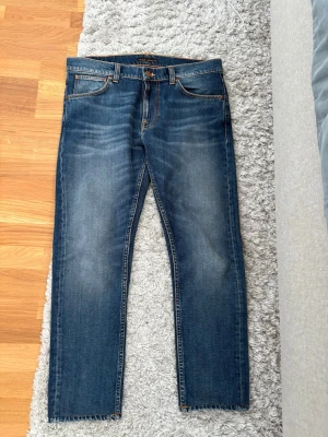 Lean Dean Nudie Jeans - Säljer ett par klassiska blå jeans från Nudie Jeans med raka ben och snygga slitningar framtill. Jeansen har fem fickor, orange sömmar och Nudie-logga på bakfickan. Tillverkade i denim med en cool tvättad look.