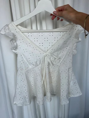 Vit omlottopp med broderie anglaise - Söt vit omlottopp med broderie anglaise och volangdetaljer vid ärmarna. Toppen har v-ringning, knyt framtill och är tillverkad i bomull. Perfekt för en fräsch och somrig look.