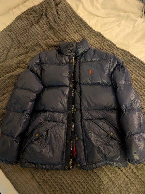 Ralph Lauren Polo dunjacka marinblå, stl M - Ralph Lauren Polo dunjacka i marinblått med ponyloggan och rött foder. OBS: lappen säger XL men det är en ungdomsstorlek (18/20) som i praktiken sitter som vuxen S/M - kolla bilderna för mått innan du köper. Fint skick. Modell: 182cm | 83kg.