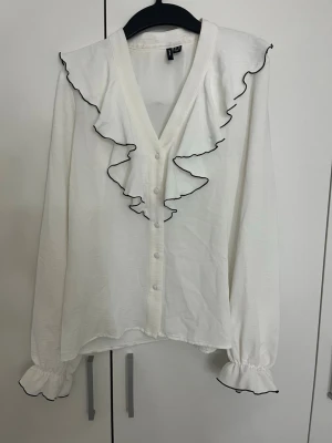 Skjorta från Vera Mode - Säljer en jättefin vit skjorta som jag köpt från Vera Moda 🤍 Den har fina volangdetaljer med svart kant runt halsen och ärmarna, vilket gör den lite extra fin. Den sitter väldigt fint på kroppen och passar perfekt både till vardags och när man vill klä upp sig lite.  Jag har endast haft på mig den en gång, så den är i väldigt fint skick. Storlek 32. Säljer för 200 kr
