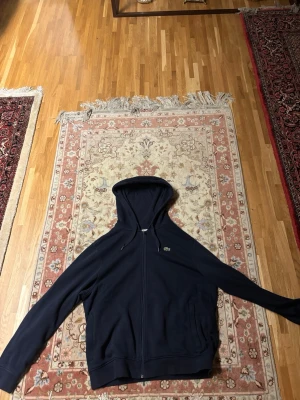 Mörkblå hoodie med dragkedja Lacoste - Snygg mörkblå hoodie från Lacoste med dragkedja framtill och klassisk krokodil-logga på bröstet. Hoodien har justerbar huva med snören och två fickor framtill. Perfekt för en avslappnad och stilren look.