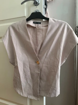 Beige linnetopp med v-ringning - Stilren beige linnetopp med v-ringning och korta ärmar. Toppen har en enkel knapp framtill och är tillverkad i ett luftigt linnematerial, perfekt för varma dagar. Passar till både jeans och kjol för en avslappnad look.