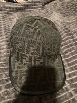 Grå keps med monogrammönster - Snygg grå keps med all-over monogrammönster i mörkare nyans. Klassisk form med böjd skärm och tygknapp på toppen. Kepsen har en stilren look och är tillverkad i tygmaterial med en kant i skinnimitation runt skärmen.
