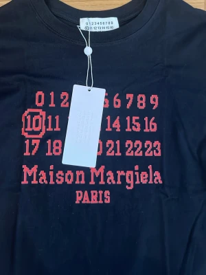 Maison Margiela svart t-shirt - Cool svart t-shirt från Maison Margiela med ikoniskt rött tryck framtill och texten 'Maison Margiela PARIS'. T-shirten har korta ärmar och är gjord i mjuk bomull. Perfekt för dig som gillar streetwear och vill sticka ut med en unik design.