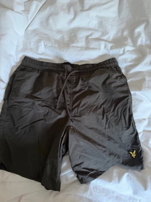 Gröna badbyxor från Lyle & Scott - Snygga gröna badbyxor från Lyle & Scott med elastisk midja och dragsko. Klassisk gul logga broderad på vänster ben. Tillverkade i snabbtorkande material med meshfoder för extra komfort. Perfekta för stranden eller poolen.